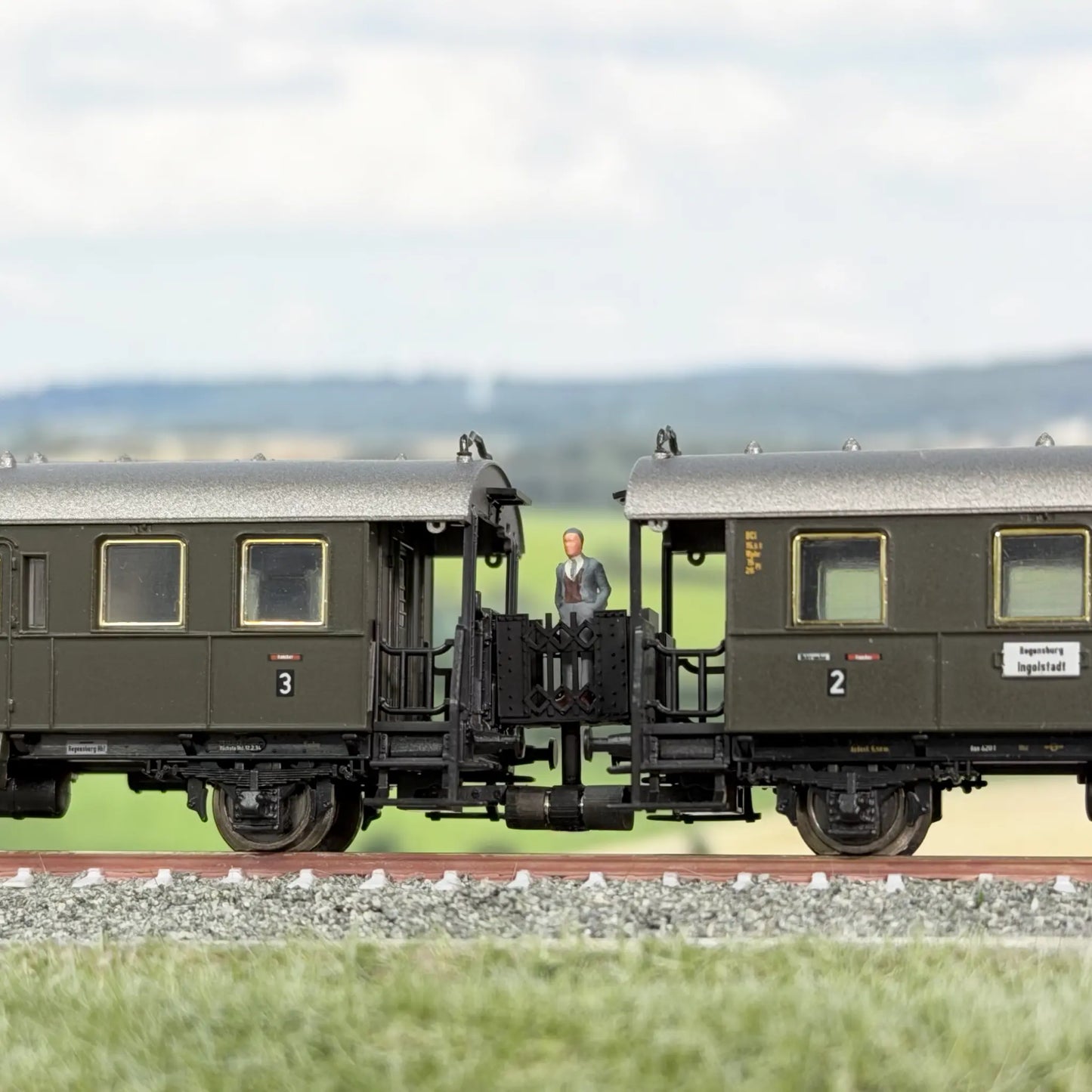 Minitrix Länderbahnwagen (Donnerbüchse), dazwischen eine Verbindungsbrücke im Minitrix Design. Auf der Brücke steht ein Fahrgast, ein Mann mit grauem Anzug und schaut sich die Landschaft an