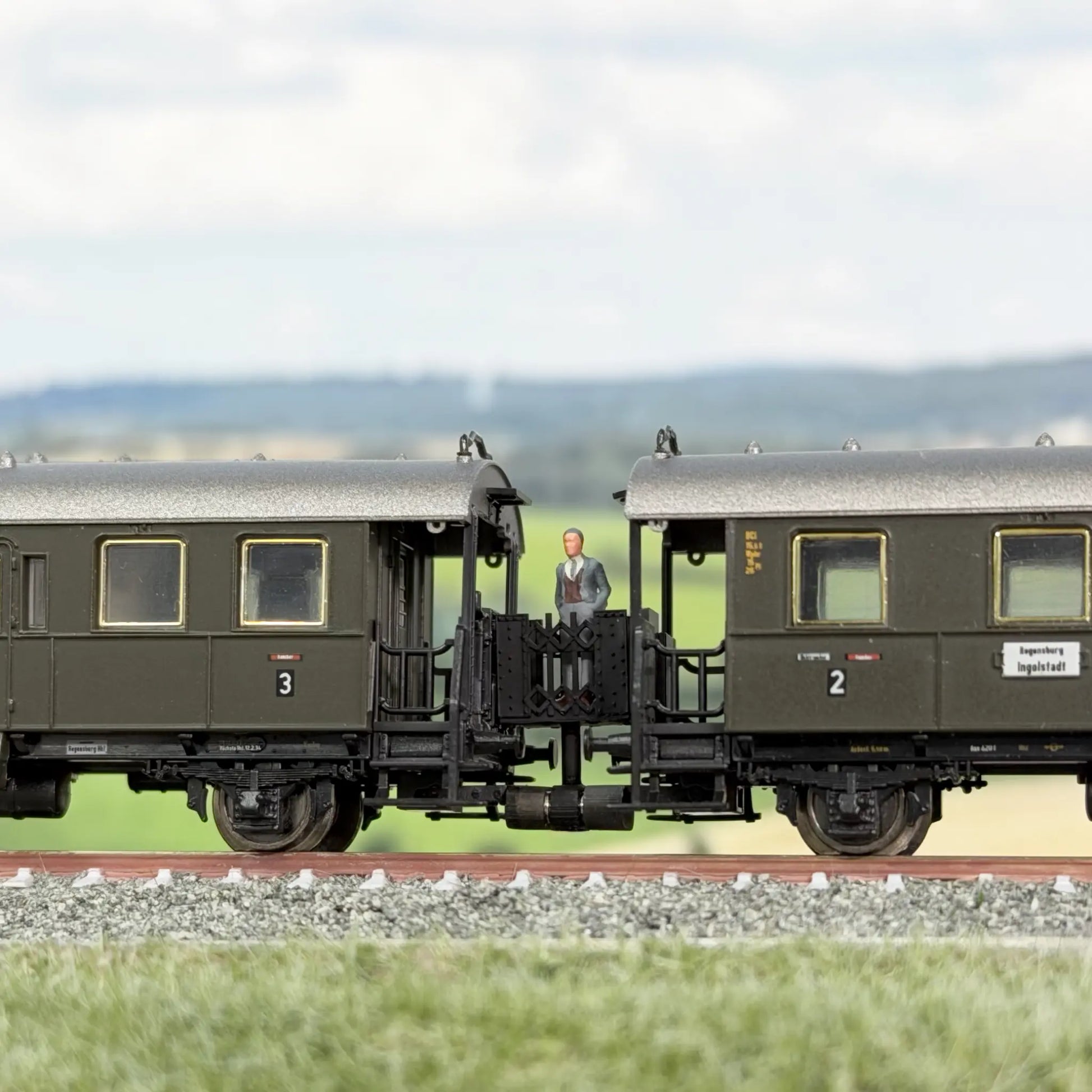 Minitrix Länderbahnwagen (Donnerbüchse), dazwischen eine Verbindungsbrücke im Minitrix Design. Auf der Brücke steht ein Fahrgast, ein Mann mit grauem Anzug und schaut sich die Landschaft an